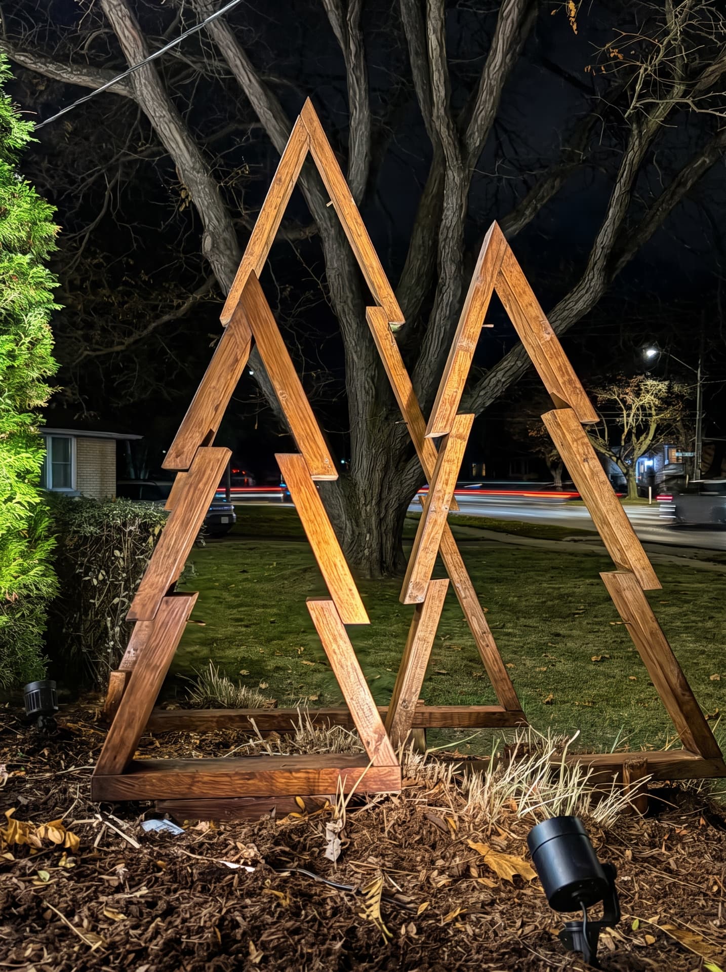 Handcrafted oversized wooden Christmas trees uplit in a front garden bed at night