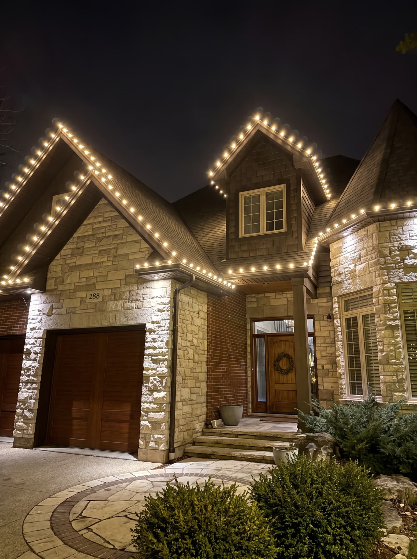 Stone and brick home with holiday lights following gables, dormers and entry peaks
